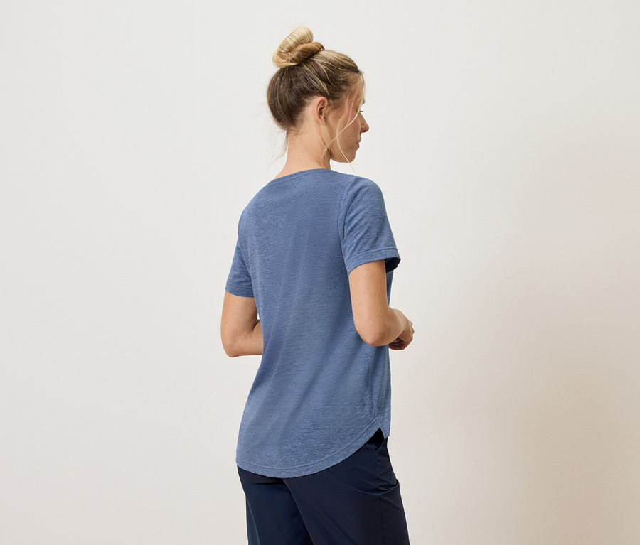 Femme vue de dos, portant un t-shirt bleu et un pantalon foncé sur un fond neutre.