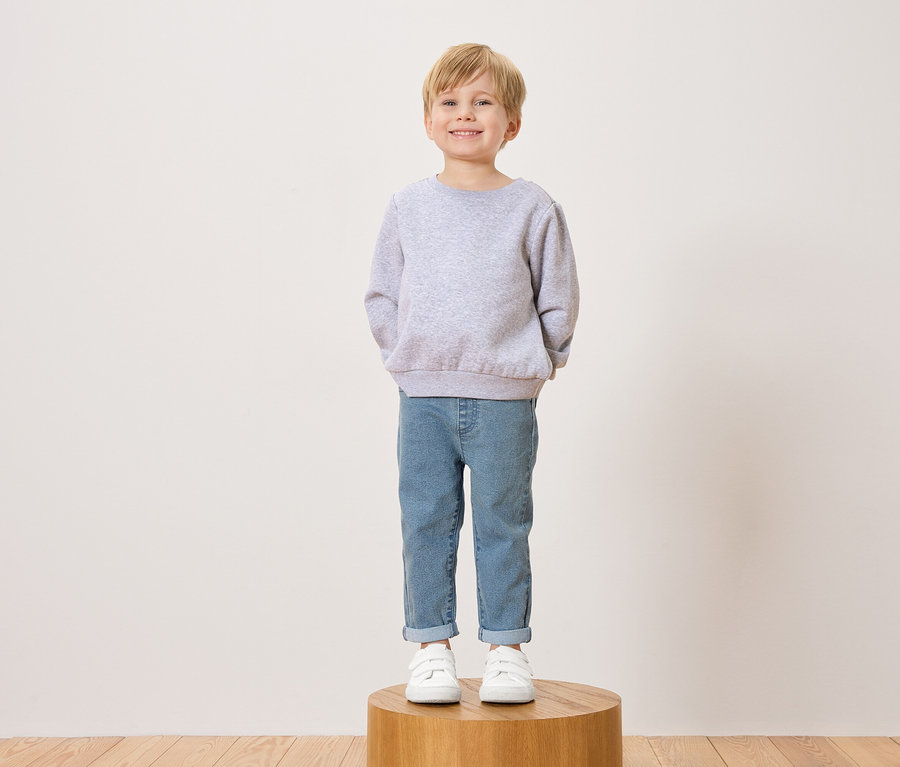 Un garçon souriant aux cheveux blonds se tient sur un tabouret en bois, vêtu d'un pull gris, d'un jean et de baskets blanches.