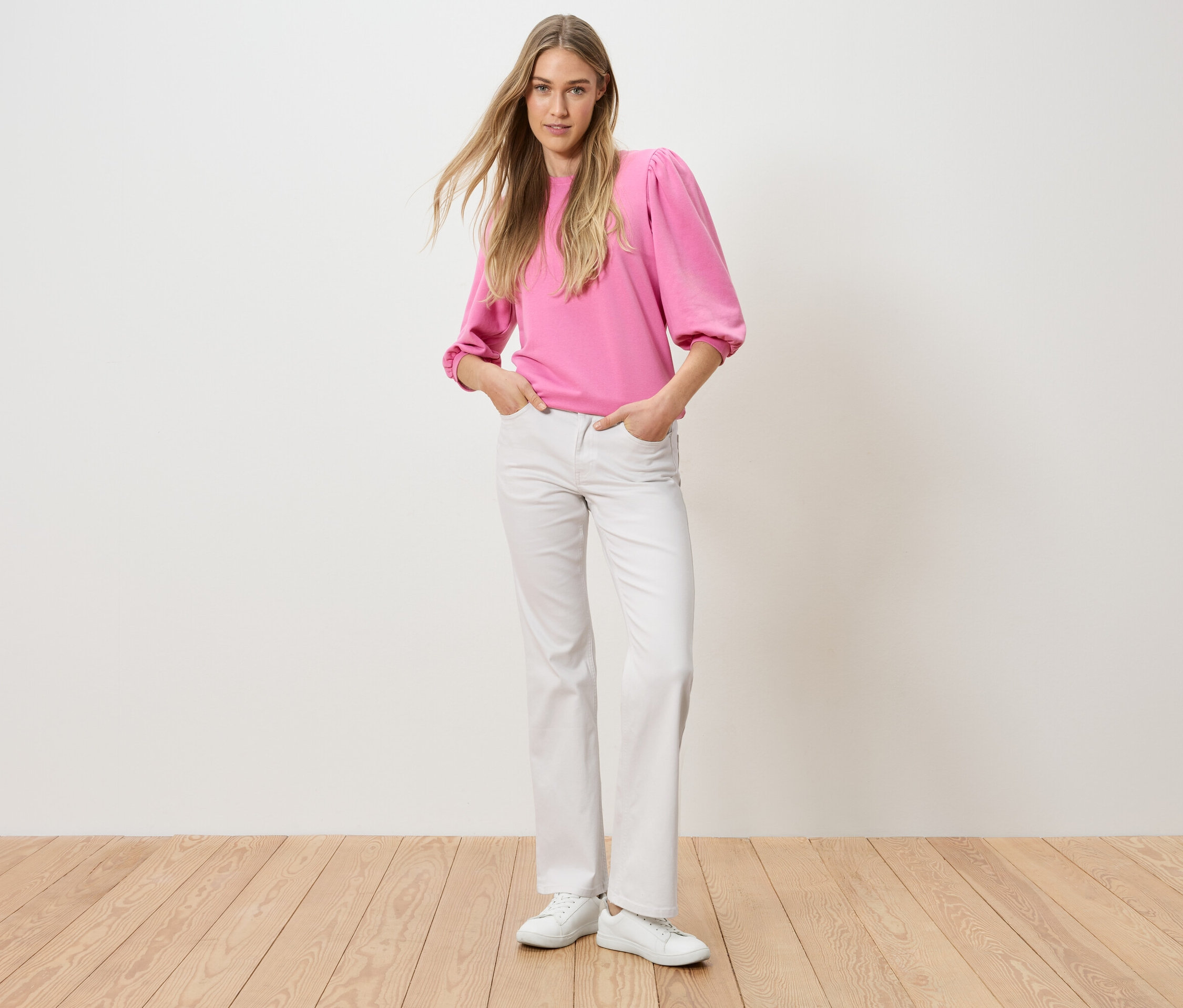 Une femme se tient debout, les mains dans les poches de son pantalon. Elle porte un haut rose et un jean blanc.