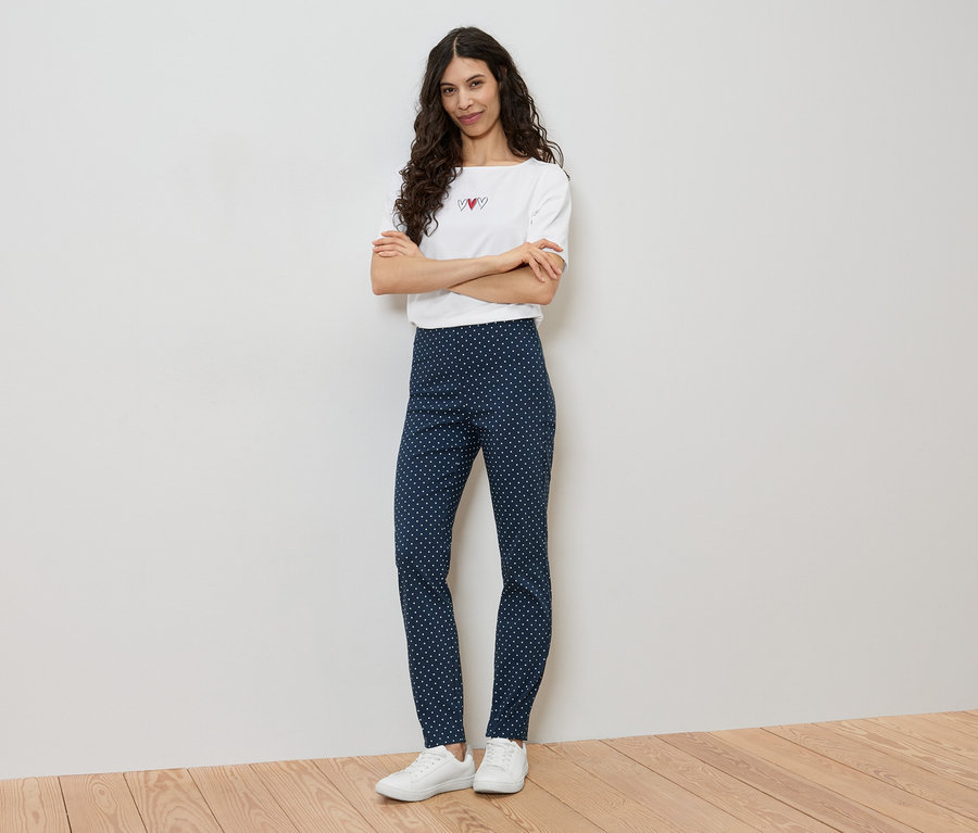Une femme se tient debout, les bras croisés, portant un t-shirt blanc avec des cœurs et un legging bleu à pois blancs.