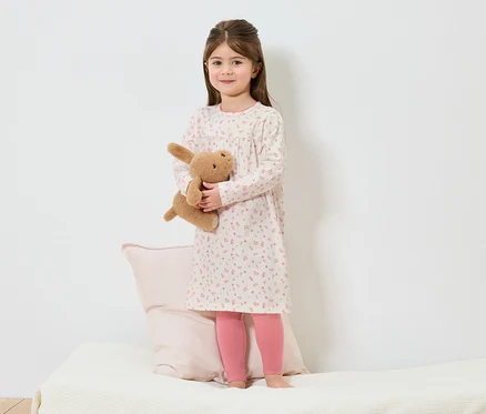 Une fille en chemise de nuit à fleurs se tient pieds nus sur un lit et tient un lapin en peluche.