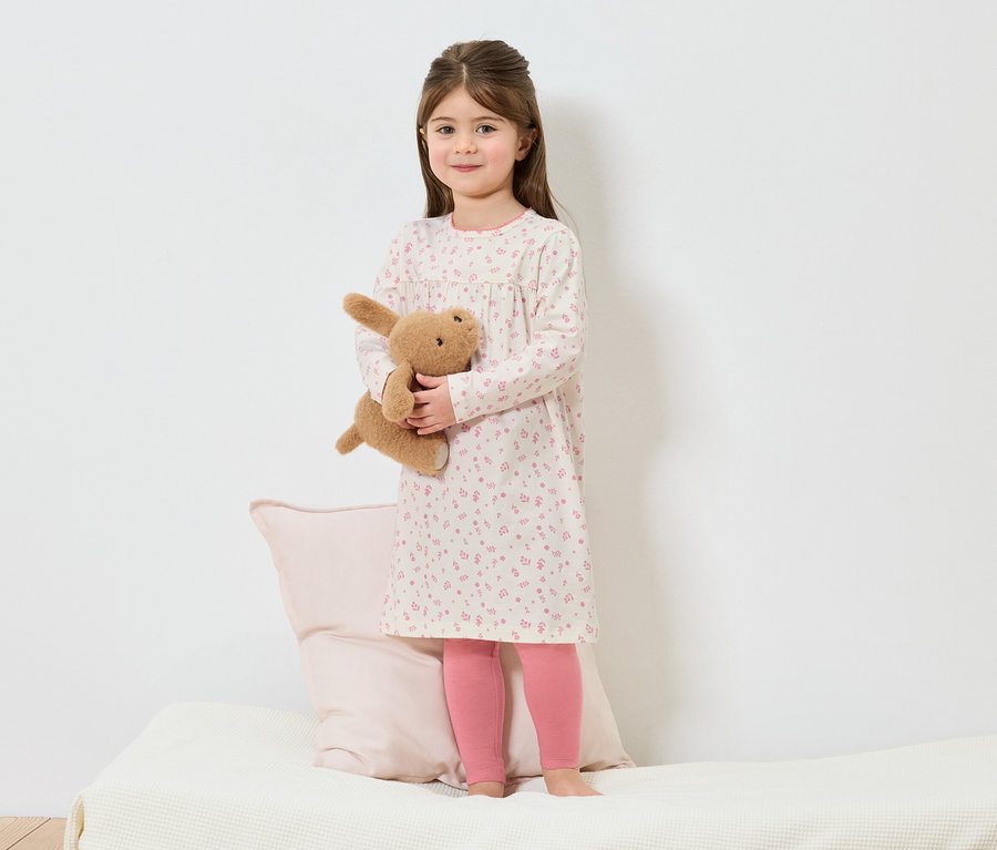 Une fille en chemise de nuit à fleurs se tient pieds nus sur un lit et tient un lapin en peluche.