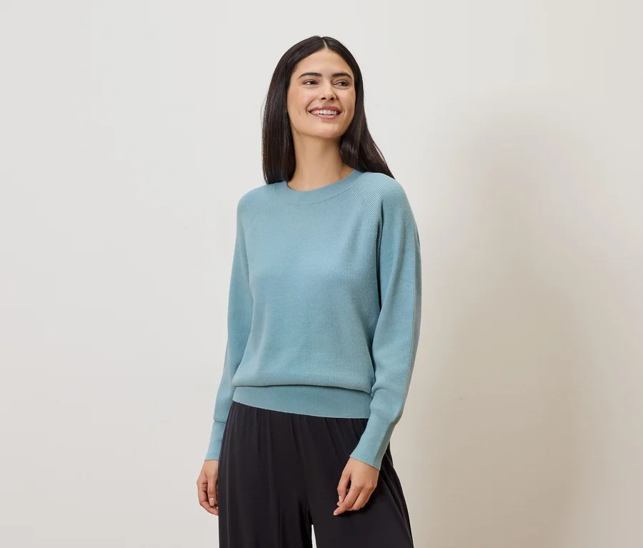 Une femme souriante aux longs cheveux foncés porte un pull bleu clair et un pantalon noir ample sur un fond clair.