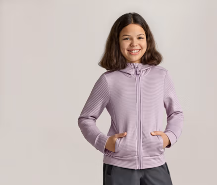 Fillette souriante, les mains dans les poches d’un sweat à capuche zippé violet et d’un pantalon gris.