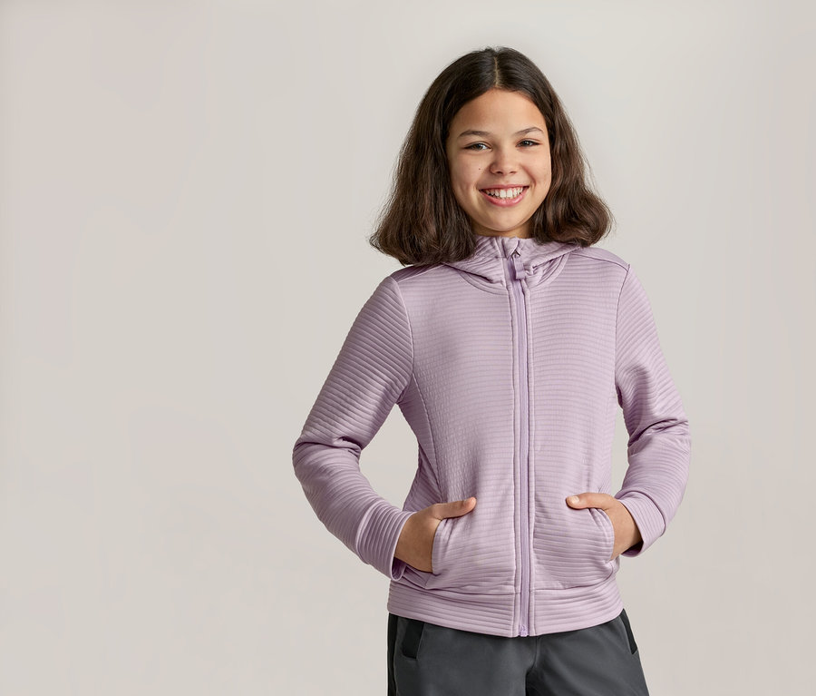 Fillette souriante, les mains dans les poches d’un sweat à capuche zippé violet et d’un pantalon gris.