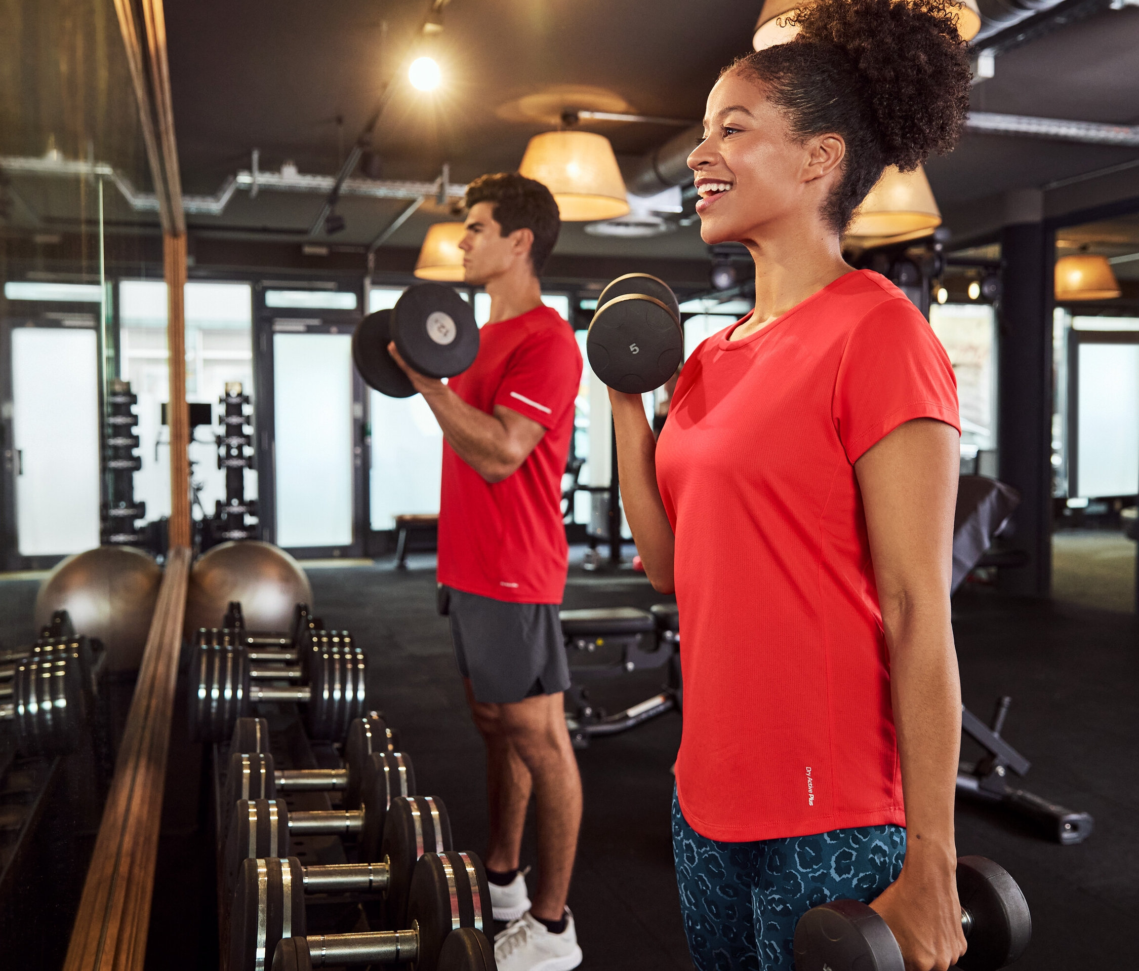 Une femme et un homme dans une salle de sport soulèvent des haltères, tous deux portant des t-shirts rouges.