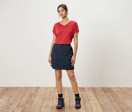 Une femme se tient devant un mur blanc, portant un t-shirt rouge, une jupe bleue et des chaussures de randonnée bleues.