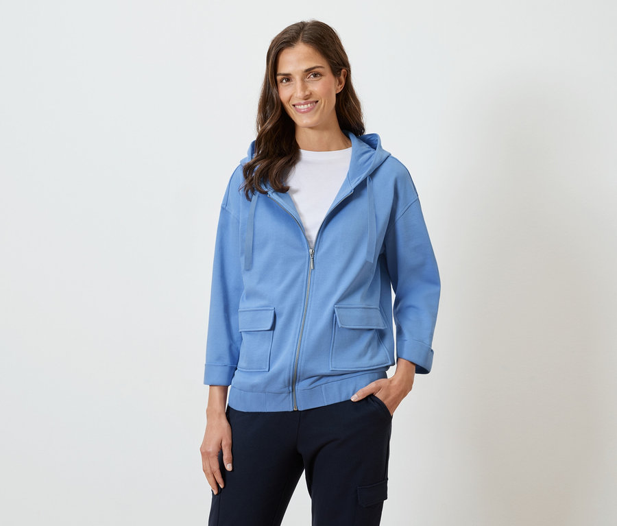 Femme aux longs cheveux bruns souriant dans un sweat à capuche bleu et un pantalon foncé.