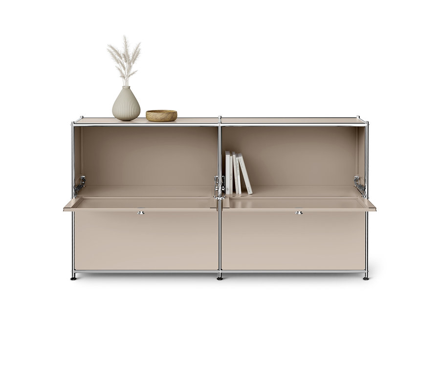 Meuble modulaire beige avec cadre chromé, comprenant deux étagères supérieures ouvertes et deux portes abattantes ouvertes en bas. Un vase avec de l'herbe de la pampa et un bol en bois sont sur l'étagère de gauche, des livres sur celle de droite.