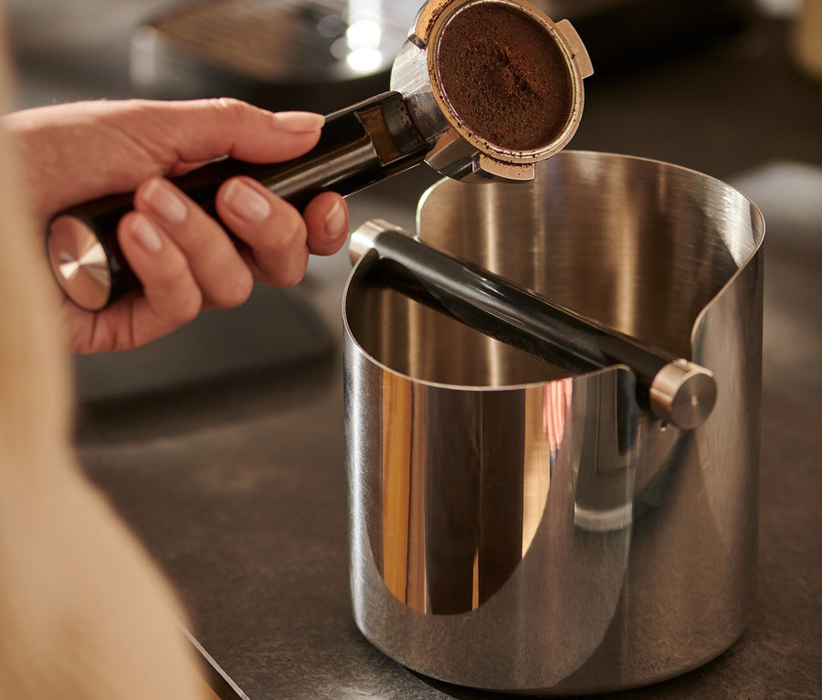 Une main tient un porte-filtre à café au-dessus d'une boîte à marc de café brillante.