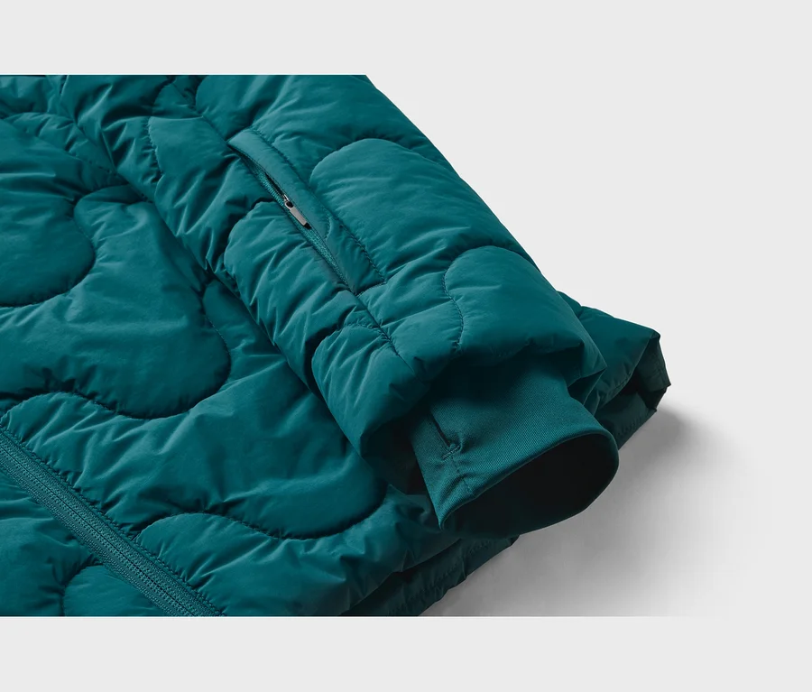 Gros plan d'une veste matelassée sarcelle, avec un motif irrégulier, une poche zippée sur la manche et un poignet côtelé.