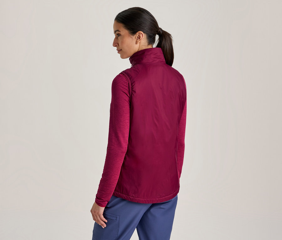 Femme vue de dos, légèrement tournée, portant un haut à manches longues magenta, un gilet magenta plus foncé et un pantalon bleu. Ses cheveux sont attachés en queue de cheval.
