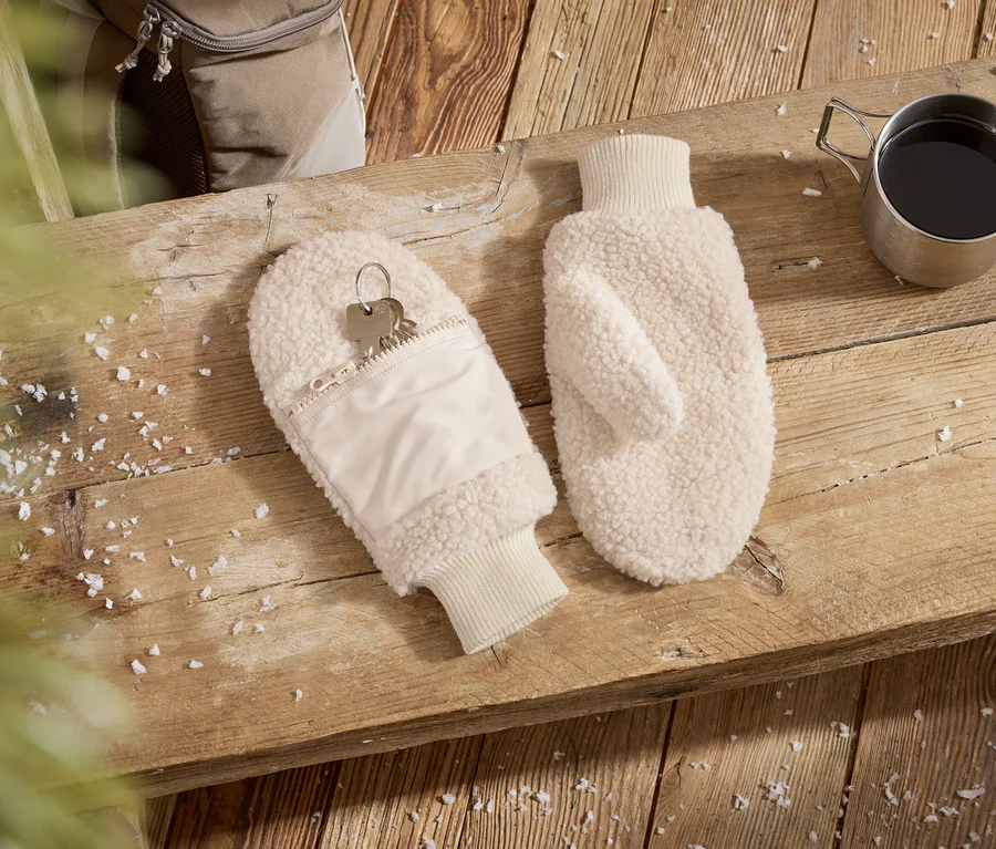 Deux moufles moelleuses de couleur crème sont posées sur une surface en bois. L'une des moufles a une poche zippée avec des clés. À côté se trouve une tasse en métal avec un liquide foncé et des flocons de neige sont éparpillés.