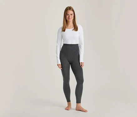 Une femme souriante est debout, pieds nus, vêtue d'un haut blanc à manches longues et d'un legging gris foncé taille haute.