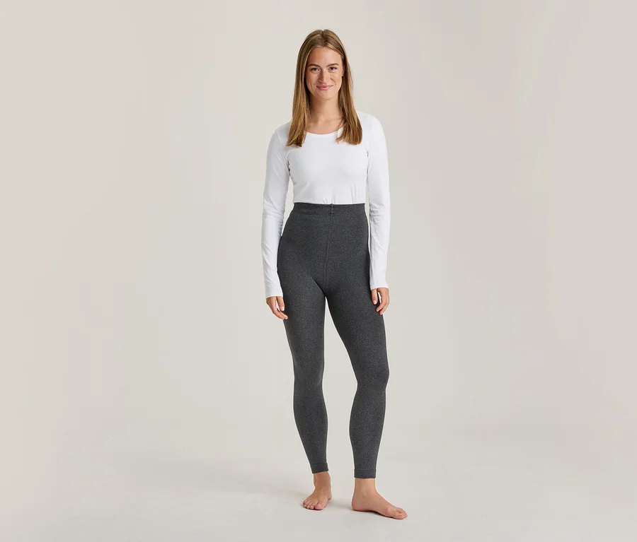 Une femme souriante est debout, pieds nus, vêtue d'un haut blanc à manches longues et d'un legging gris foncé taille haute.