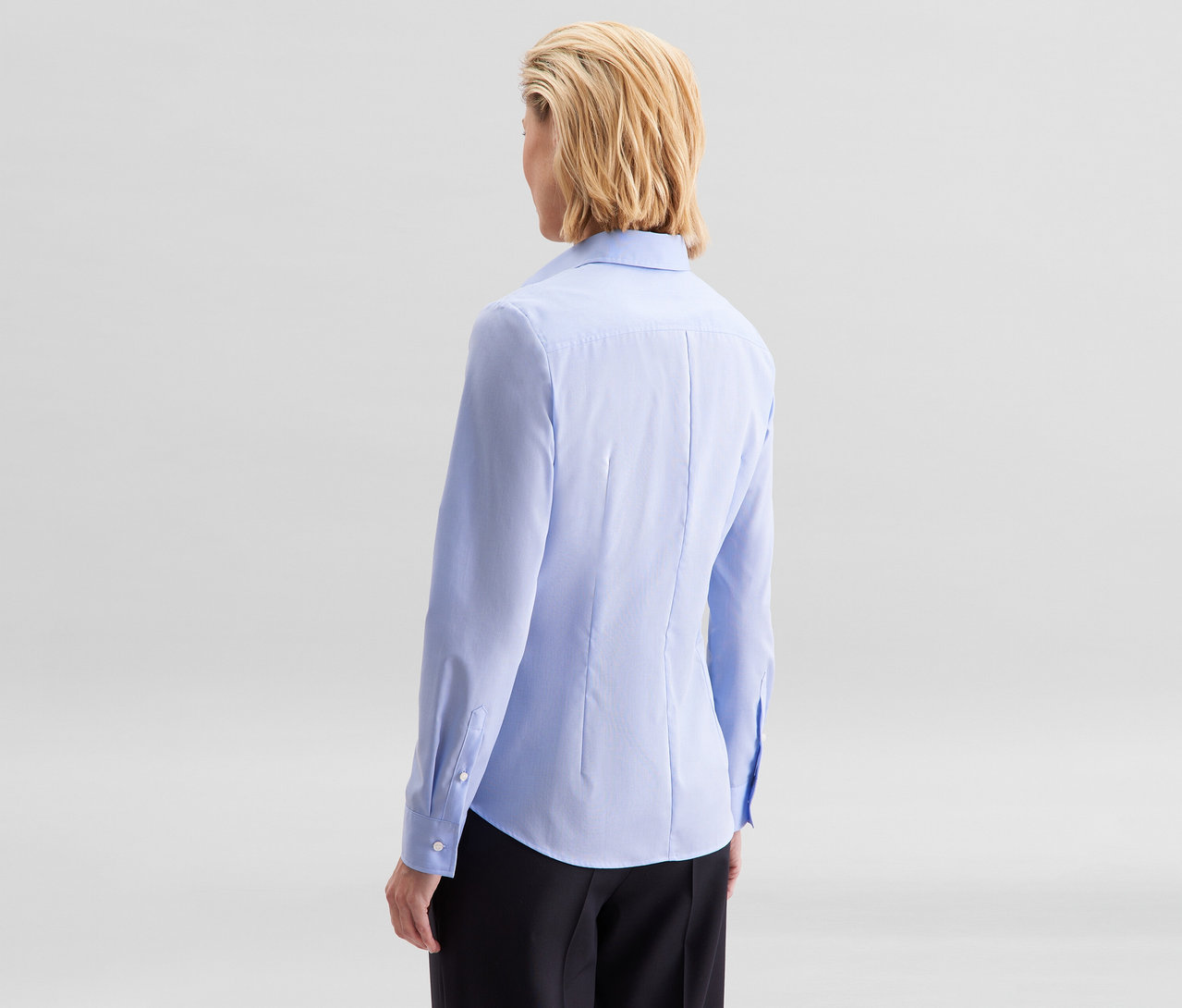 Femme de dos, aux cheveux blonds courts, portant un chemisier Slim Fit non iron Seidensticker bleu clair et un pantalon noir.