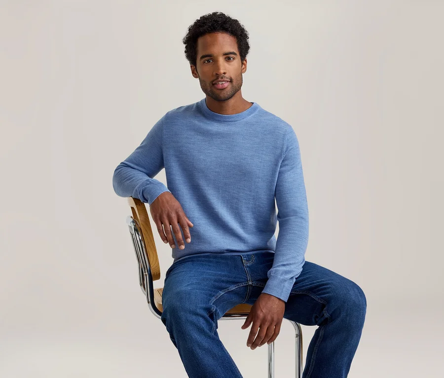 Un homme aux cheveux foncés et bouclés, portant un pull bleu clair à manches longues et un jean bleu, est assis sur une chaise et regarde devant lui.