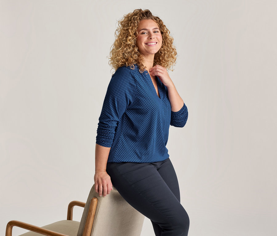 Femme souriante aux cheveux blonds bouclés, vêtue d'un chemisier bleu à motifs et d'un pantalon foncé, assise de côté sur le dossier d'une chaise claire.
