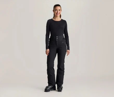 Une femme souriante se tient de face, vêtue d'un haut noir à manches longues, d'un pantalon de ski noir taille haute avec boutons et poches zippées, et de chaussures de ski noires.