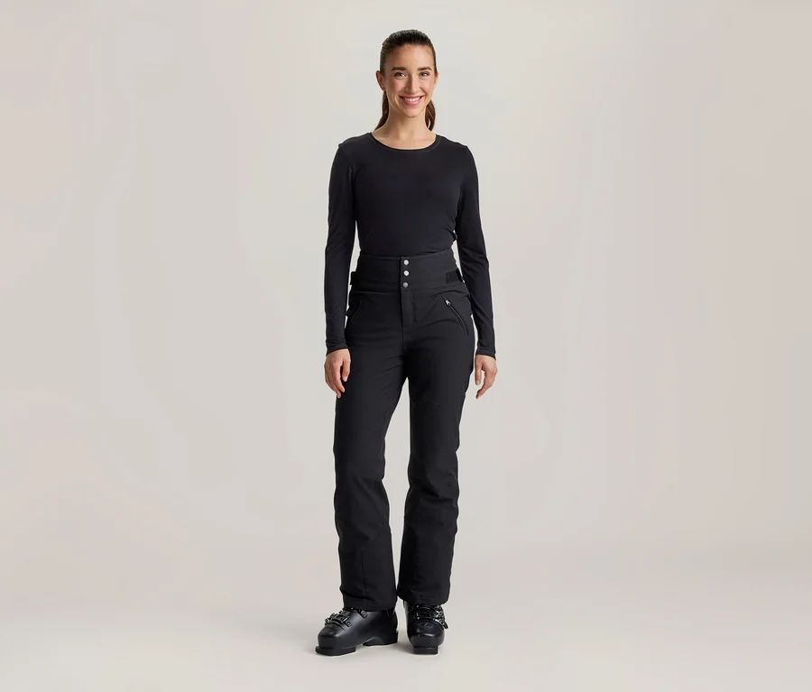 Une femme souriante se tient de face, vêtue d'un haut noir à manches longues, d'un pantalon de ski noir taille haute avec boutons et poches zippées, et de chaussures de ski noires.