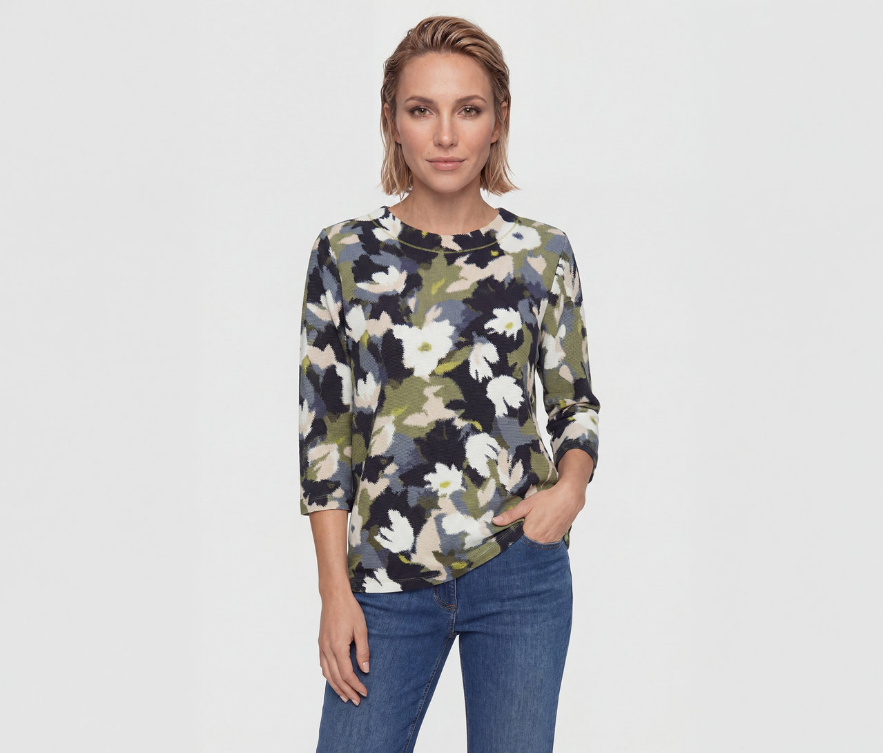 Femme aux cheveux blonds courts portant un haut floral (noir, blanc, vert, beige, gris) et un jean bleu, une main dans la poche.