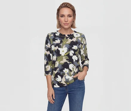 Femme aux cheveux blonds courts portant un haut floral (noir, blanc, vert, beige, gris) et un jean bleu, une main dans la poche.