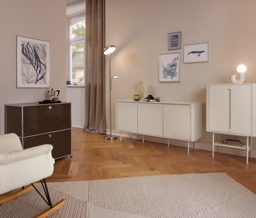 Un salon confortable avec des murs clairs et un parquet en bois. Au premier plan, un fauteuil à bascule crème est placé à côté d'une armoire marron foncé avec une cafetière moka. Un lampadaire se trouve entre un long buffet blanc et une armoire blanche plus haute. Diverses œuvres d'art encadrées ornent les murs, et un tapis clair couvre une partie du sol.