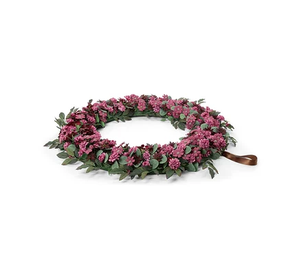 Une couronne florale ronde avec de petites fleurs roses et violettes et des feuilles vertes, avec une boucle de ruban marron sur un côté, sur un fond blanc.