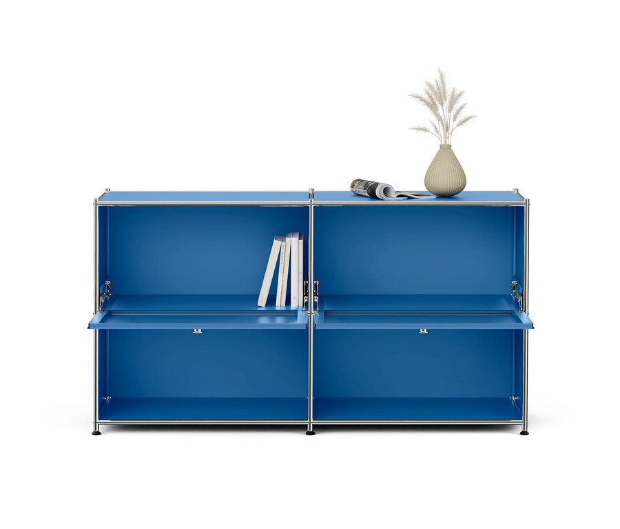 Meuble modulaire bleu avec cadre chromé, composé de deux éléments à deux compartiments chacun. Les portes abattantes inférieures sont légèrement ouvertes. Des livres sont rangés dans le compartiment supérieur gauche, et un journal roulé ainsi qu'un vase avec des herbes séchées sont posés sur le dessus droit.