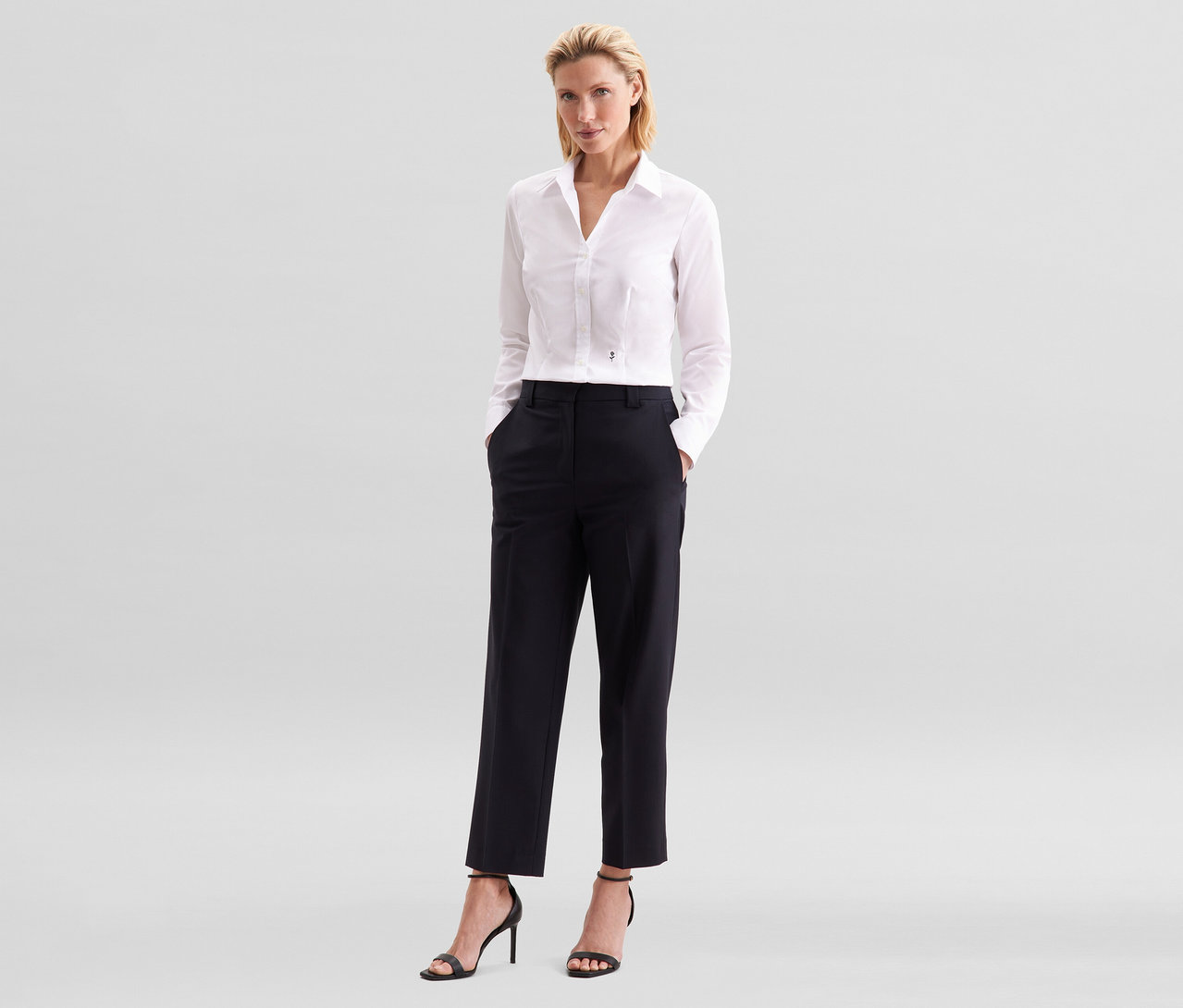 Une femme aux cheveux blonds se tient les mains dans les poches de son pantalon noir. Elle porte un chemisier Slim Fit non iron Seidensticker blanc et des sandales noires à talons hauts.