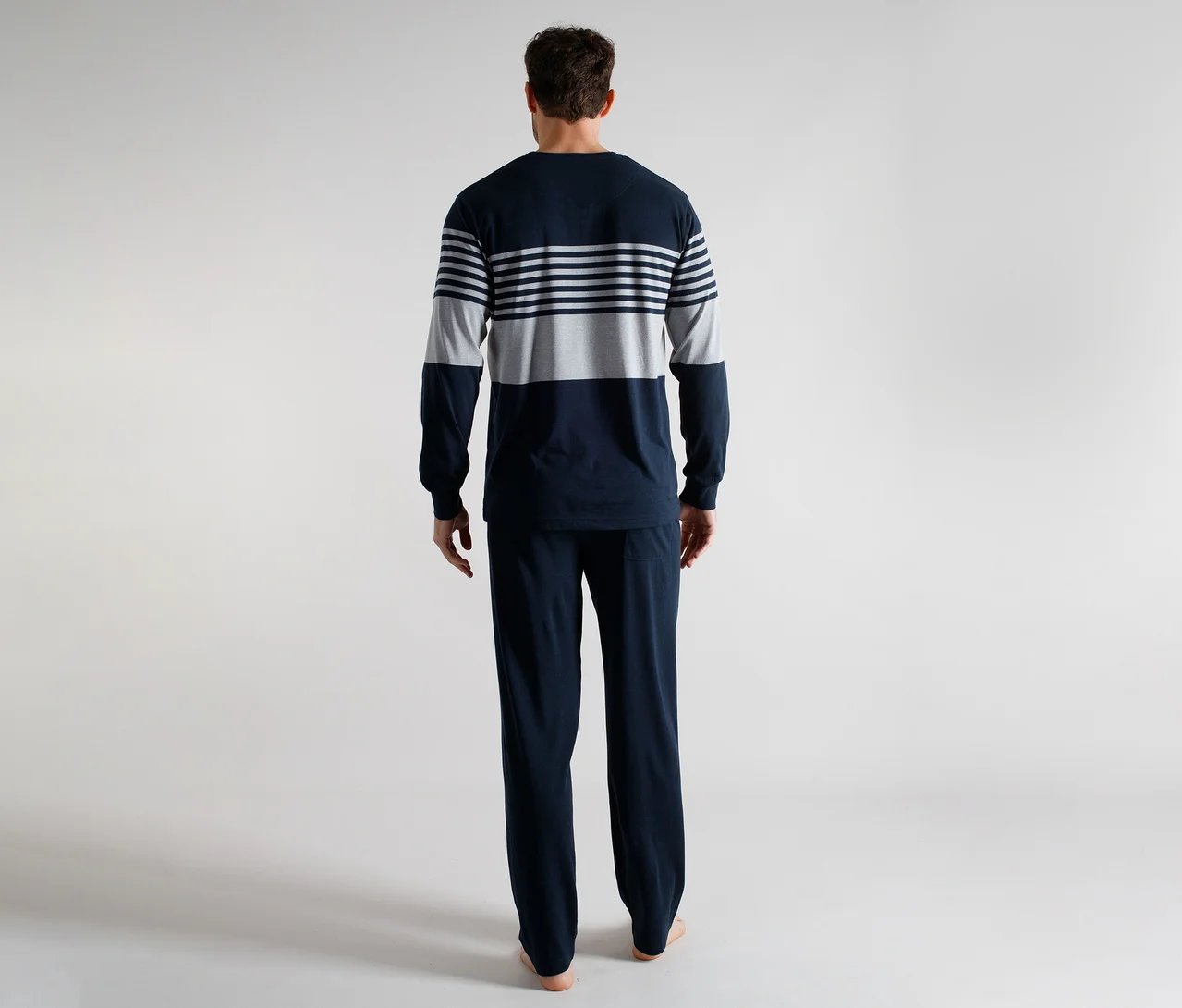 Homme de dos portant un pyjama à manches longues rayé bleu marine et gris et un pantalon de pyjama bleu marine uni, debout pieds nus.