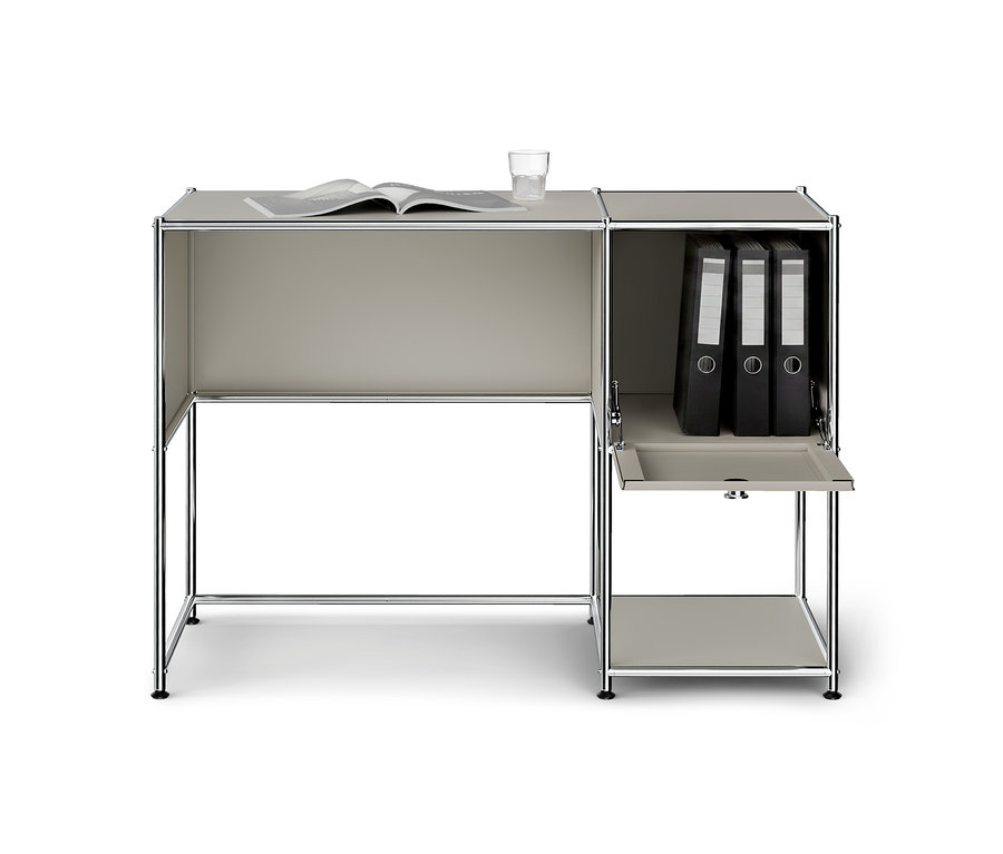 Bureau modulaire gris clair avec pieds chromés. Un livre ouvert et un verre d'eau sont posés sur le bureau. L'unité de rangement attenante à droite contient trois classeurs noirs, et une porte abattante inférieure est ouverte, révélant une étagère.