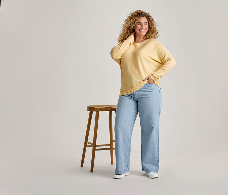 Une femme souriante aux cheveux bouclés, vêtue d'un haut jaune à manches longues et d'un jean large bleu clair, se tient à côté d'un tabouret en bois.