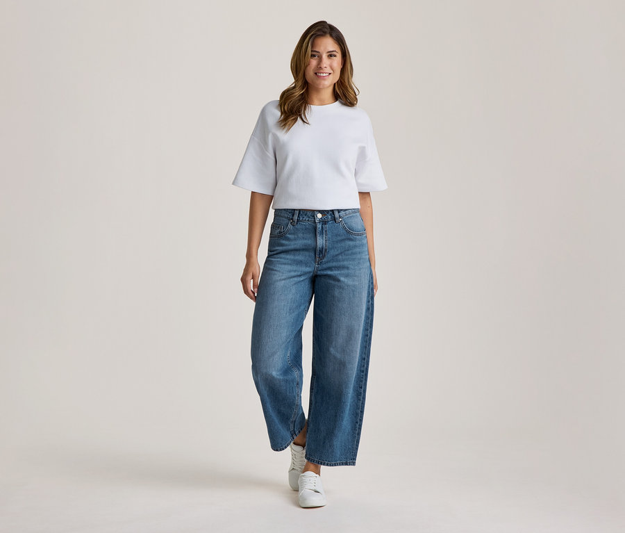 Une femme souriante se tient de face, vêtue d'un t-shirt blanc à manches larges, d'un jean bleu large et de baskets blanches.