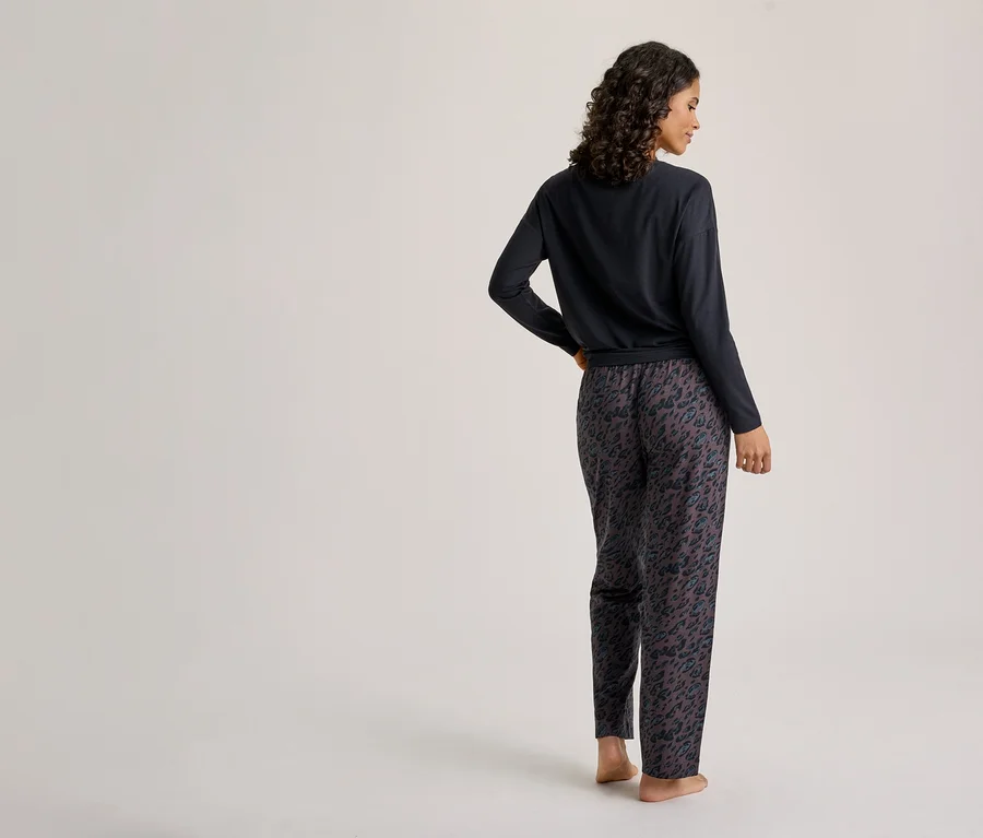 Une femme est debout, de dos, regardant légèrement par-dessus son épaule droite. Elle porte un haut foncé à manches longues et un pantalon de pyjama foncé à motif animalier. Ses pieds sont nus.