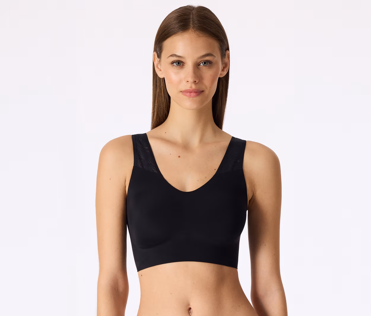 Femme aux longs cheveux bruns portant une brassière de sport noire avec un col en V et des détails en maille sur les bretelles, sur fond blanc.