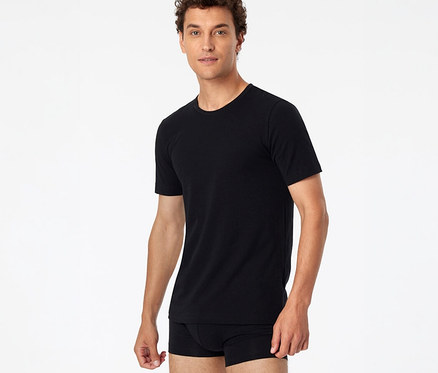 Homme aux cheveux bruns bouclés portant un t-shirt SCHIESSER noir et un boxer noir, posant devant un fond blanc.