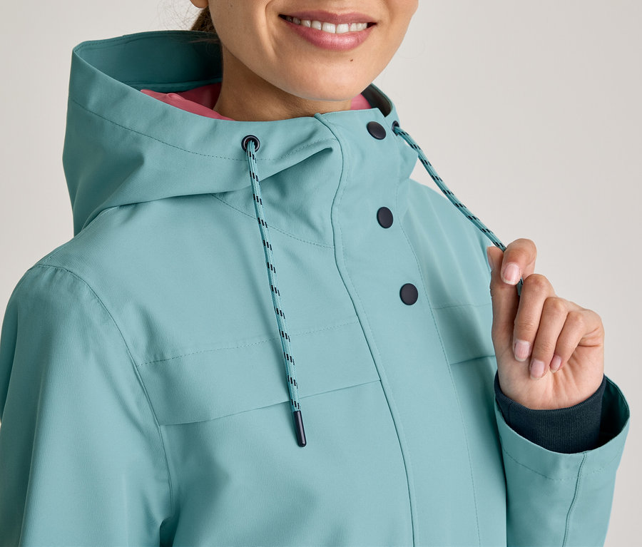 Personne souriante portant une veste à capuche turquoise avec des boutons foncés et une doublure rose. Sa main tire l'un des cordons de la capuche.
