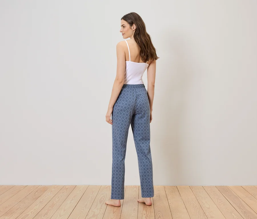 Une femme en débardeur blanc et pantalon bleu à motifs se tient sur un plancher en bois devant un mur blanc.