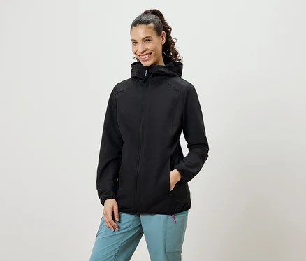Une femme souriante avec une queue de cheval porte une veste softshell noire et un pantalon bleu.