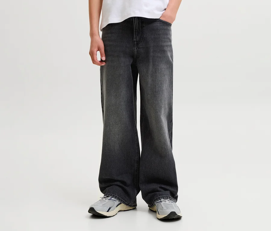 Personne portant un jean large gris foncé, un t-shirt blanc et des baskets gris clair, une main dans la poche.