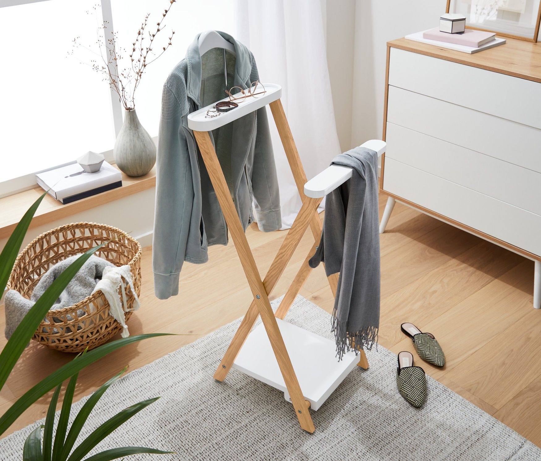 Pièce lumineuse avec un portant en bois sur lequel sont suspendus une veste bleu clair, une écharpe grise et des lunettes. En arrière-plan, une commode, une fenêtre et des décorations.