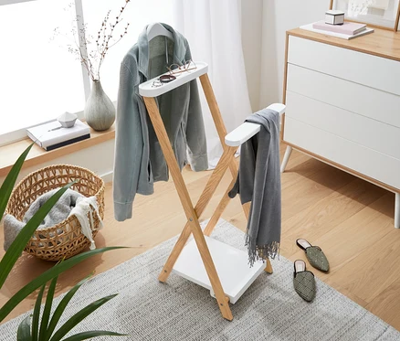 Pièce lumineuse avec un portant en bois sur lequel sont suspendus une veste bleu clair, une écharpe grise et des lunettes. En arrière-plan, une commode, une fenêtre et des décorations.