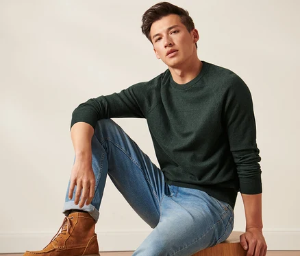 Un homme est assis et porte un pull vert foncé à col rond, un jean et des bottes marron.