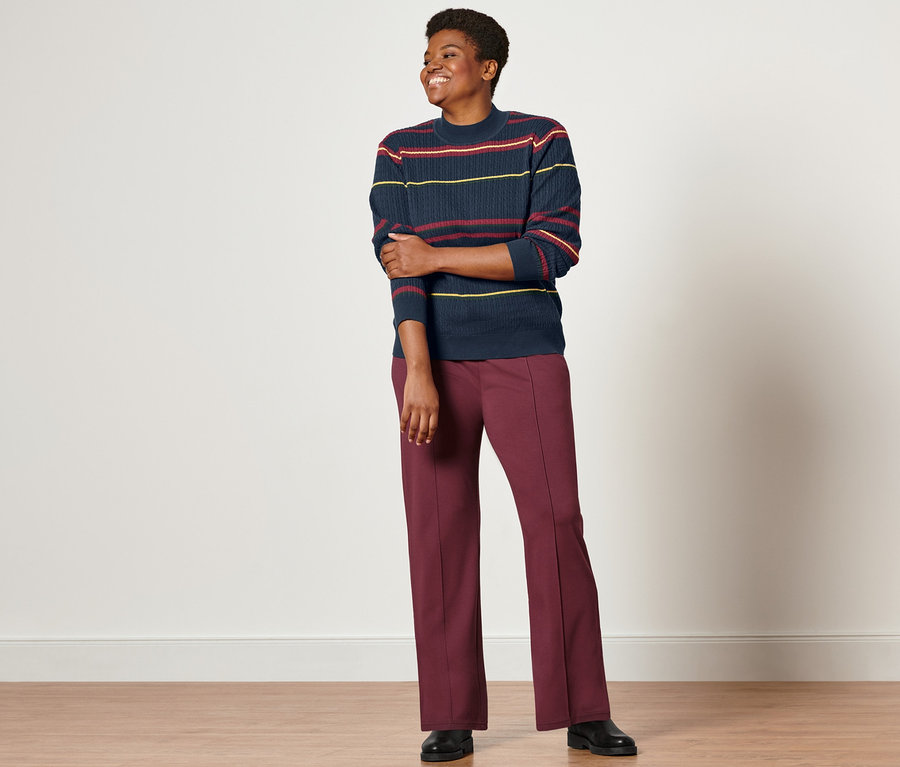 Une femme debout porte un pull en maille fine multicolore avec torsades, un pantalon de jogging avec nervure et des bottines en cuir.