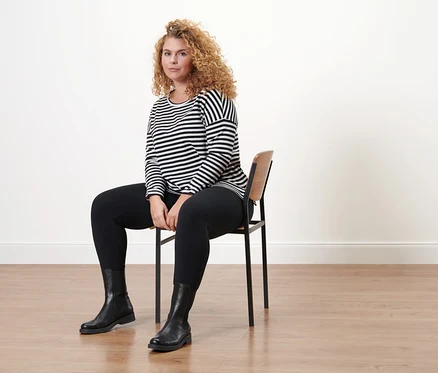 Une femme est assise sur une chaise et porte des bottines en cuir et un t-shirt manches longues à rayures.