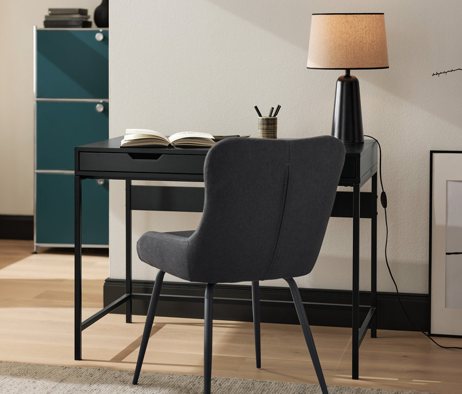Une chaise rembourrée anthracite est placée à un bureau noir. Sur le bureau se trouvent une lampe de table noire avec abat-jour en tissu et un livre ouvert.