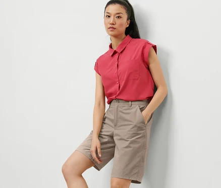 Une femme appuyée contre un mur porte un short fonctionnel en softshell taupe et une chemise rouge.