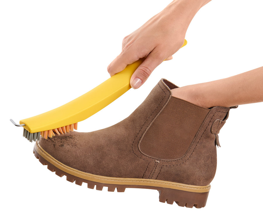 Une main nettoie une chaussure marron en daim avec une brosse à chaussures 3 en 1.