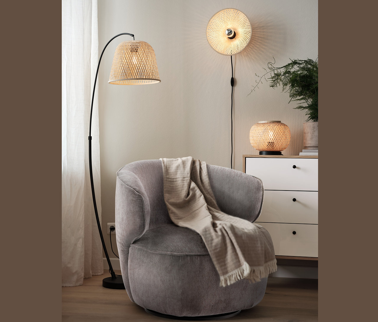 Un fauteuil pivotant gris avec une couverture est placé à côté d'une lampe de recourbée avec abat-jour en bambou. Une lampe murale avec abat-jour en bambou est sur le mur et une lampe de table avec abat-jour est sur la commode.